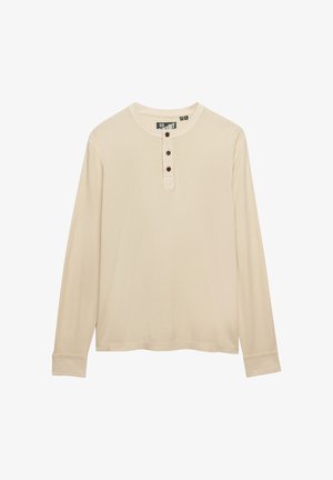 Beżowa koszula henley z długim rękawem, trzema ciemnymi guzikami na listwie i okrągłym dekoltem.