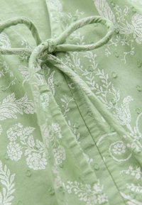 Vihreä kuvioitu kangas, jossa on valkoisia kukkakuvioita ja teksturoituja pisteitä. Ominaisuuksiin kuuluu sidottu yksityiskohta pääntiessä ja ommellut saumat.