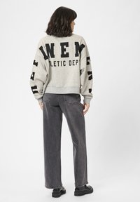 Donna con capelli scuri alla spalla, che indossa una felpa oversize grigia chiara con lettering nero e jeans grigi scuri, in piedi con le spalle rivolte verso di noi.