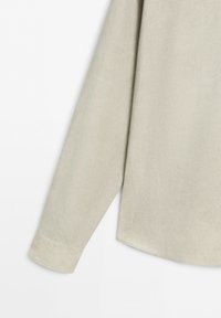 Manica lunga beige di camicia con polsino e cucitura laterale, mostrata su sfondo bianco.