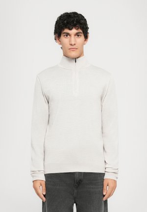 Man med lockigt hår som bär en ljusgrå dragkedjetröja med hög hals och mörka jeans, stående mot en helt vit bakgrund.