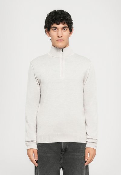 Mann mit lockigem Haar, der einen hellgrauen Reißverschluss-Pullover mit Stehkragen und dunklen Jeans trägt und vor einem schlichten weißen Hintergrund steht.