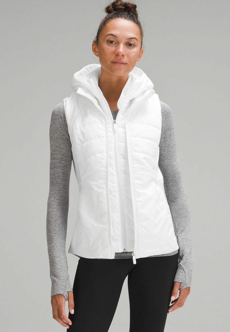 Gilet imbottito bianco con cappuccio e chiusura zip, caratterizzato da un design trapuntato. Indossato sopra una maglietta a maniche lunghe grigia, con tessuto leggero e forma aderente.