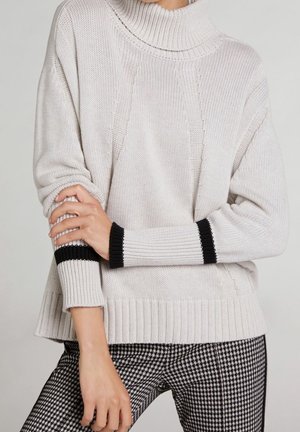 Pull en tricot gris clair avec un grand col, présentant des poignets à rayures noires et une coupe décontractée, associé à un pantalon en pied-de-poule noir et blanc.