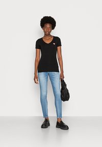 T-shirt nera a maniche corte con scollo a V e logo rosso, abbinata a jeans skinny blu chiaro e scarpe chunky nere; la modella tiene una borsa nera.