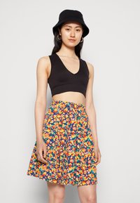 Top curto preto, saia plissada com um padrão floral vibrante, com flores laranjas, rosas e verdes sobre um fundo azul escuro. Chapéu tipo balde.