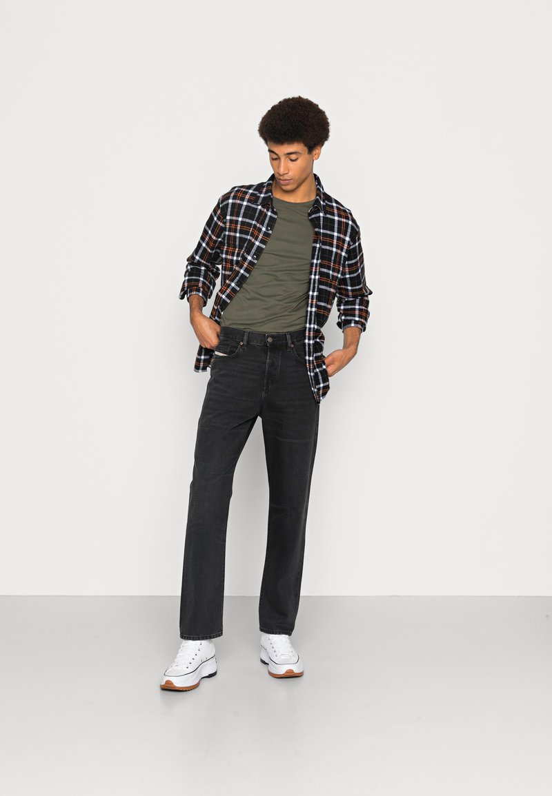 Zwarte geruite shirt over een groene T-shirt, gecombineerd met zwarte jeans en witte sneakers. Het shirt heeft rode en blauwe accenten.