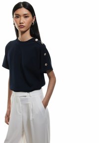 Marinblå kortärmad topp med dekorativa guldknapputstickare på axeln, tillsammans med högmidjade vita skräddarsydda byxor. Slät textur.