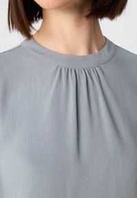 Hellgraue Bluse mit rundem Halsauschnitt, strukturiertem Stoff und dezenten Falten unterhalb des Halsausschnitts. Glatte Oberfläche ohne sichtbare Beschläge.