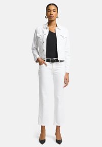 Giacca di denim bianco sopra un top nero, jeans bianchi a gamba larga e tacchi neri. Cintura nera con fibbia ovale. La modella sta in piedi con una mano sui fianchi.