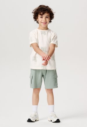 T-shirt beige chiaro con stampa del logo grigia, abbinata a bermuda cargo color menta della lunghezza del ginocchio. L'outfit è completato da calze bianche e sneaker.