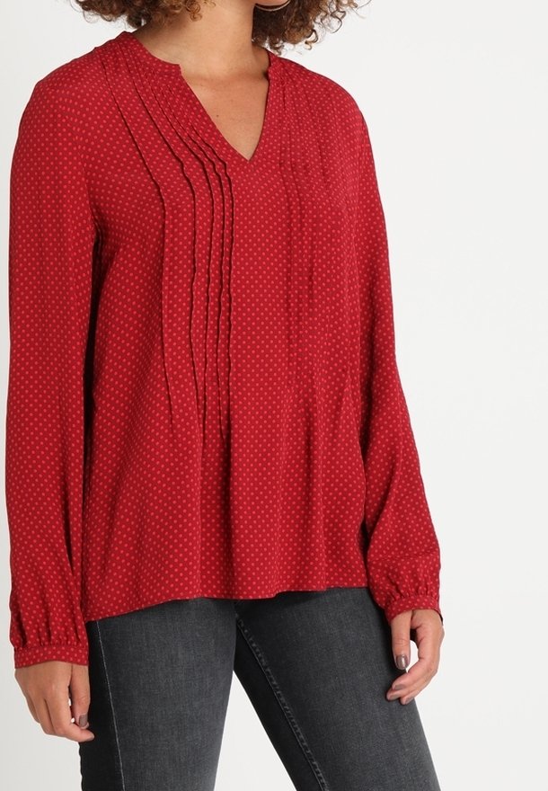 Rode blouse met V-hals, geplooide voorkant en lange mouwen. Heeft een stippenpatroon en een losse pasvorm. Het materiaal lijkt zacht en gestructureerd.