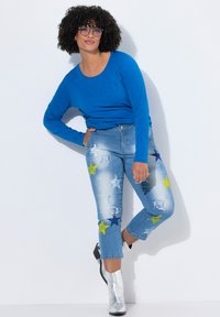 Top azul de manga larga combinado con jeans azul claro que presentan parches de estrellas multicolores y detalles desgastados; el modelo lleva botines plateados.