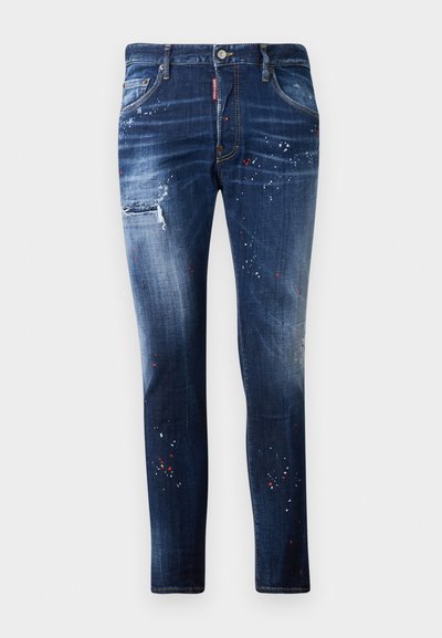 Jeans de ganga azul escuro com um corte justo, remendos desgastados e detalhes de tinta salpicada em branco e vermelho. Design clássico de cinco bolsos.