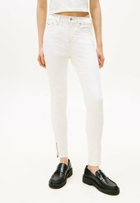 Jeans skinny blancos con un detalle de cremallera en los tobillos. El material es suave y tiene un ligero estiramiento. Combinados con zapatos de charol negros.