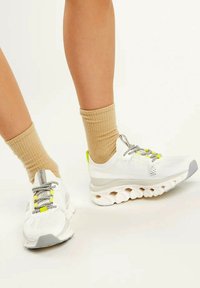 Scarpe sportive bianche con accenti grigi, tomaia in rete testurizzata, lacci riflettenti, dettagli giallo neon e una suola imbottita e segmentata.