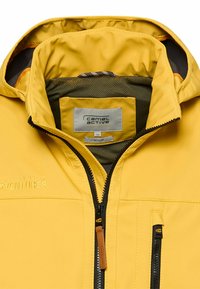 Gelbe Outdoorjacke mit Frontreißverschluss und Reißverschluss an der Brusttasche, ausgestattet mit Innenfutter aus Mesh und Camel Active Markenetikett im Krageninneren.