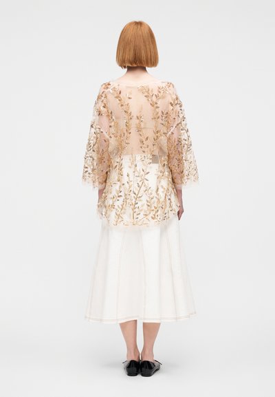 Αέρινη, κεντητή μπλούζα με σχέδιο σε χρυσό λουλούδι, συνδυασμένη με μια δομημένη, λευκή φούστα με ορατές ραφές. Μαύρα μπαλαρίνια ολοκληρώνουν το σύνολο.