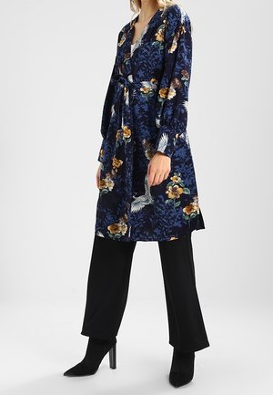 Femme portant une robe portefeuille fleurie bleu marine avec ceinture sur un pantalon noir large et des bottines à talons noires.