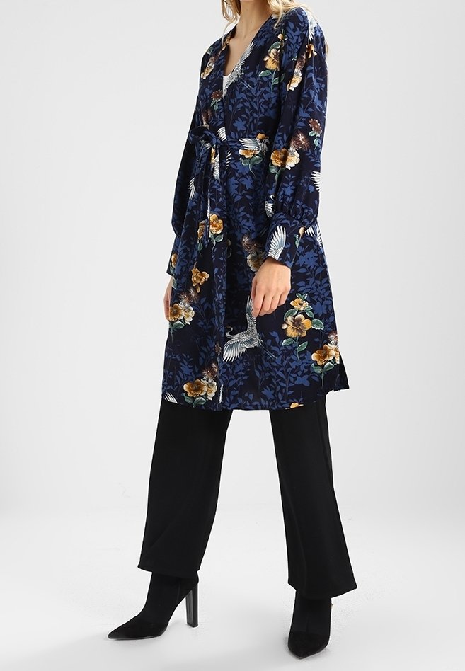 Femme portant une robe portefeuille fleurie bleu marine avec ceinture sur un pantalon noir large et des bottines à talons noires.