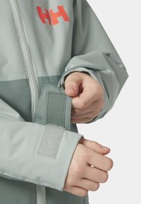 Světlé šedé vodotěsné bundě s oranžovým logem. Obsahuje zip vpředu, nastavitelné rukávy se suchým zipem a kontrastní tmavě šedou panel.