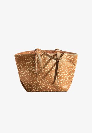 Jonge vrouw in een wit hemdje en beige broek houdt een grote bruine tas met patroon vast tegen een effen lichte achtergrond.