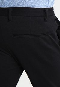 Pantalon noir à texture lisse, doté d'une poche arrière unique et d'une ceinture plate. Le tissu semble durable et bien coupé.