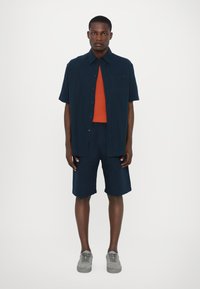 Ensemble de chemise à manches courtes et de shorts bleu marine, superposé à une chemise orange, fabriqué dans un tissu léger au toucher doux. Des baskets grises complètent le look.