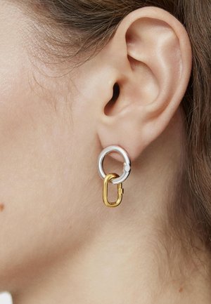 Pendientes circulares de plata y oro con anillos entrelazados y un diseño de broche, resaltando acabados metálicos contrastantes y una textura suave.