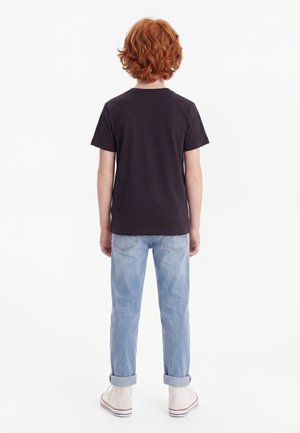 Barn med krøllet rødt hår står med ryggen til iført en sort t-shirt, lyseblå opsmøgede jeans og hvide høje sneakers.