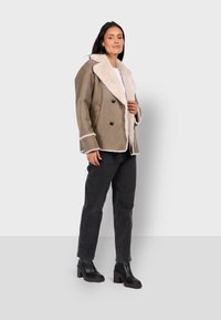 Giacca in suede marrone doppiopetto con colletto in pelliccia sintetica bianca, dotata di bottoni neri e cuciture a contrasto. Indossata con jeans neri e stivali.