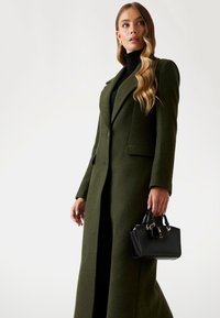 Palton lung, croit pe corp, din lâna verde închis, cu guler, asortat cu o geantă neagră cu accesorii aurii și un detaliu de breloc.