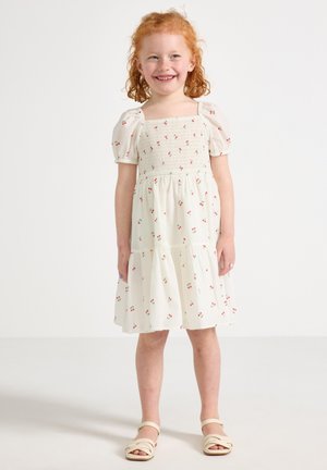Witte jurk met korte pofmouwen en een gesmockte bovenkant, met een kersenpatroon. Het heeft een gelaagde rok en is knie-lang.