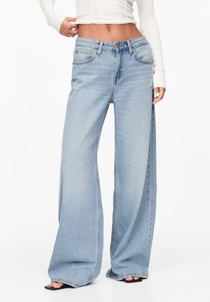 Femme portant un jean large bleu clair et un pull court blanc côtelé, debout avec une main près de la taille.
