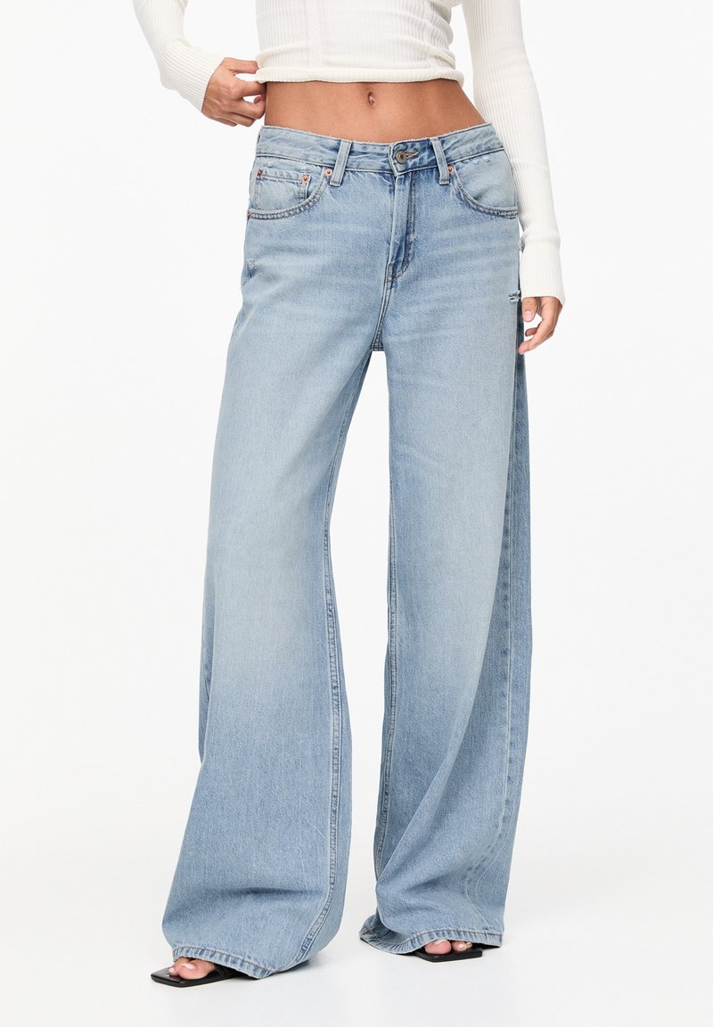 Femme portant un jean large bleu clair et un pull court blanc côtelé, debout avec une main près de la taille.