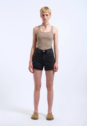 Ung person med kort blondt hår iført en beige tanktop, sorte denimshorts, brune slip-on sko og et brunt slips som tilbehør, stående.
