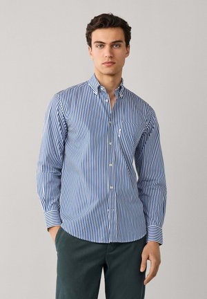 Joven con camisa de botones a rayas verticales azules y blancas y pantalones verde oscuro, de pie frente a un fondo claro y liso.