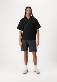 Camisa negra de manga corta con botones y textura acanalada, combinada con pantalones cortos de mezclilla gris oscuro y zapatillas beige con acentos blancos.