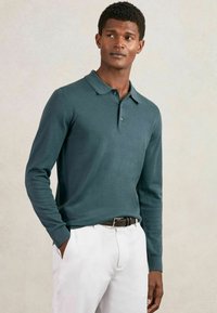 Polo cu mânecă lungă dintr-un material tricotat de culoare teal, cu guler clasic, placket cu trei butoane și manșete ribbate, asortat cu pantaloni albi și un curea maro.