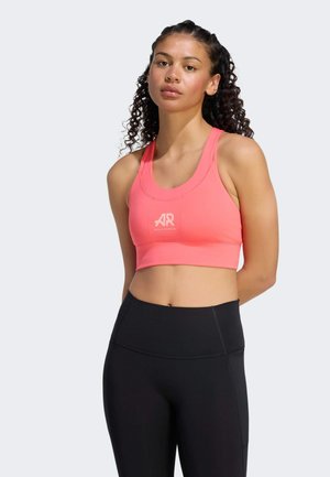 Donna con capelli ricci che indossa un reggiseno sportivo rosa e leggings neri a vita alta, in piedi davanti a uno sfondo chiaro e uniforme.