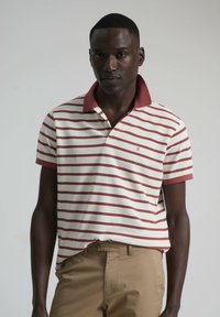 Polo de rayas en color crema y burdeos, con un cuello burdeos sólido. Mangas cortas y un diseño de corte ajustado, combinado con pantalones beige.
