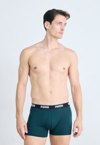 Bărbat fără tricou cu piele deschisă și păr închis la culoare, purtând boxeri Puma de culoare verde închis, privind în lateral pe un fundal deschis.