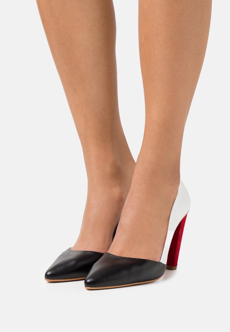 Femme portant des chaussures à talons hauts noires, blanches et rouges à bouts pointus sur un fond blanc uni.