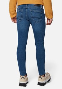 Jeans ajustados en denim azul oscuro, con dos bolsillos traseros con costuras delineadas, combinados con zapatillas beige y un suéter amarillo.