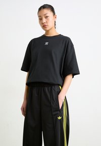 Black cotton t-shirt with loose fit and crew neckline, featuring a small white logo. Paired with black pants, highlighted by yellow side stripes.