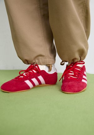 Röda mockaskor för sport med vita ränder, platt gummisula och röda snören. Bärs med beige byxor, placerade på en grön yta.