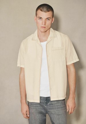 Young man with short hair wearing a cream short-sleeve button-up shirt over a white T-shirt and faded blue jeans, standing against a plain background.