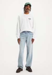 Witte overhemd met lange mouwen en zwart Levi's logo, lichtblauwe rechte jeans en donkerbruine schoenen. Groene pet voegt een contrasterende kleur toe.