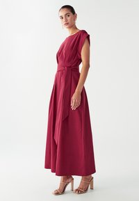 Burgundy-mekko, jossa on virtaava siluetti, lyhyet hihat ja vyöyksikkö vyötäröllä. Sileä kangas; yhdistetty beigeihin korkokenkiin.