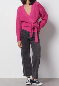 Ljusröd stickad wrap-kofta med knytbälte, ihop med mörkgrå raka jeans och svart-vita sneakers.